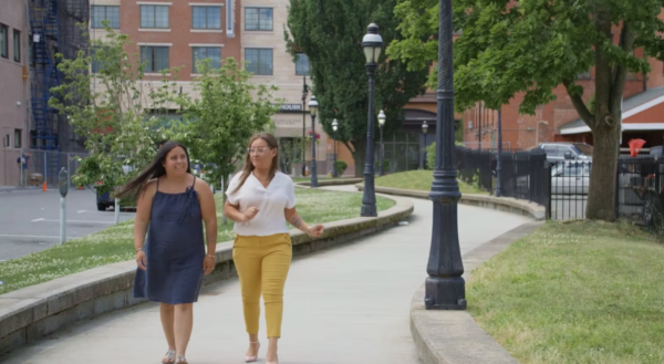 Two women walking on a path beside a grassy area with trees, buildings, and street lamps in the background.